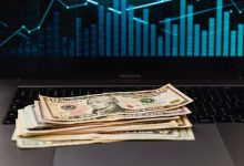 Cédulas de dólar sobre um laptop, com um gráfico financeiro ao fundo.