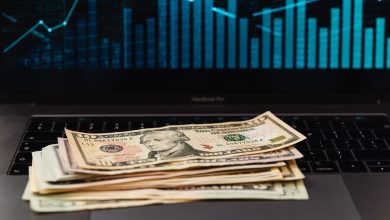 Cédulas de dólar sobre um laptop, com um gráfico financeiro ao fundo.