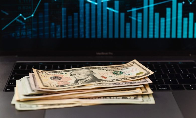 Cédulas de dólar sobre um laptop, com um gráfico financeiro ao fundo.