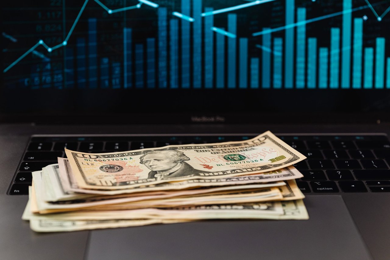 Cédulas de dólar sobre um laptop, com um gráfico financeiro ao fundo.