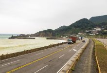 Caminhões trafegam por uma rodovia costeira cercada por montanhas e mar.