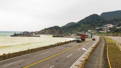 Caminhões trafegam por uma rodovia costeira cercada por montanhas e mar.