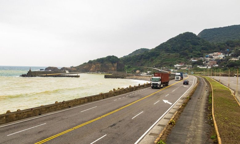 Caminhões trafegam por uma rodovia costeira cercada por montanhas e mar.